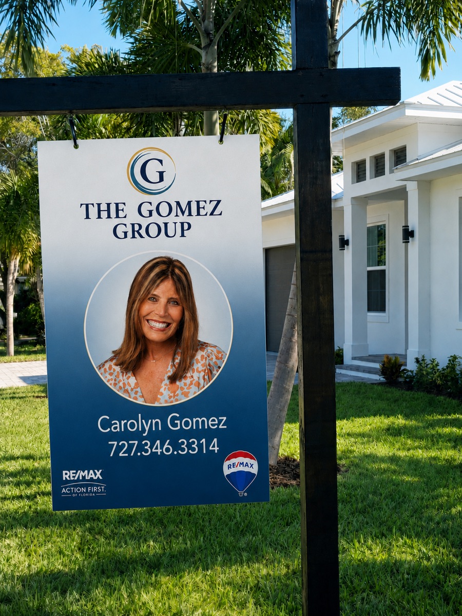 Hanging post yard sign in front of a coastal home: the full-color Storm Navy and Soft Brass seal lockup over a Storm Navy gradient panel, Carolyn's circular headshot, name, phone, and RE/MAX Action First co-brand
